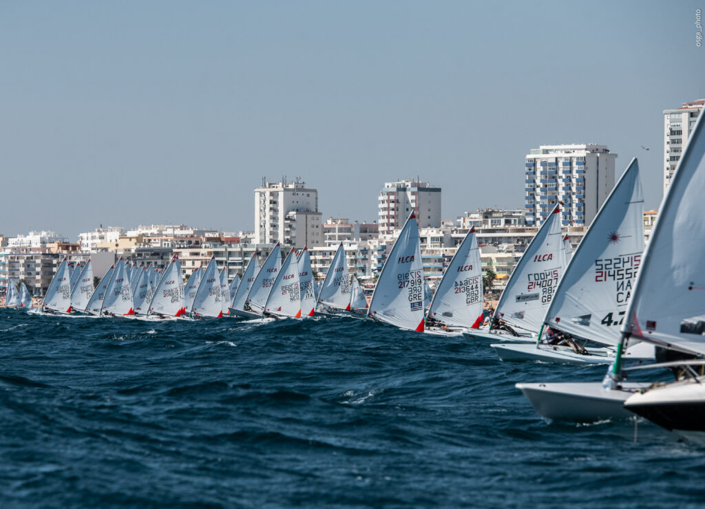 Multiple sailboats line up on the water near the coast of Vilamoura, with city buildings visible in the background, during a sailing competition.