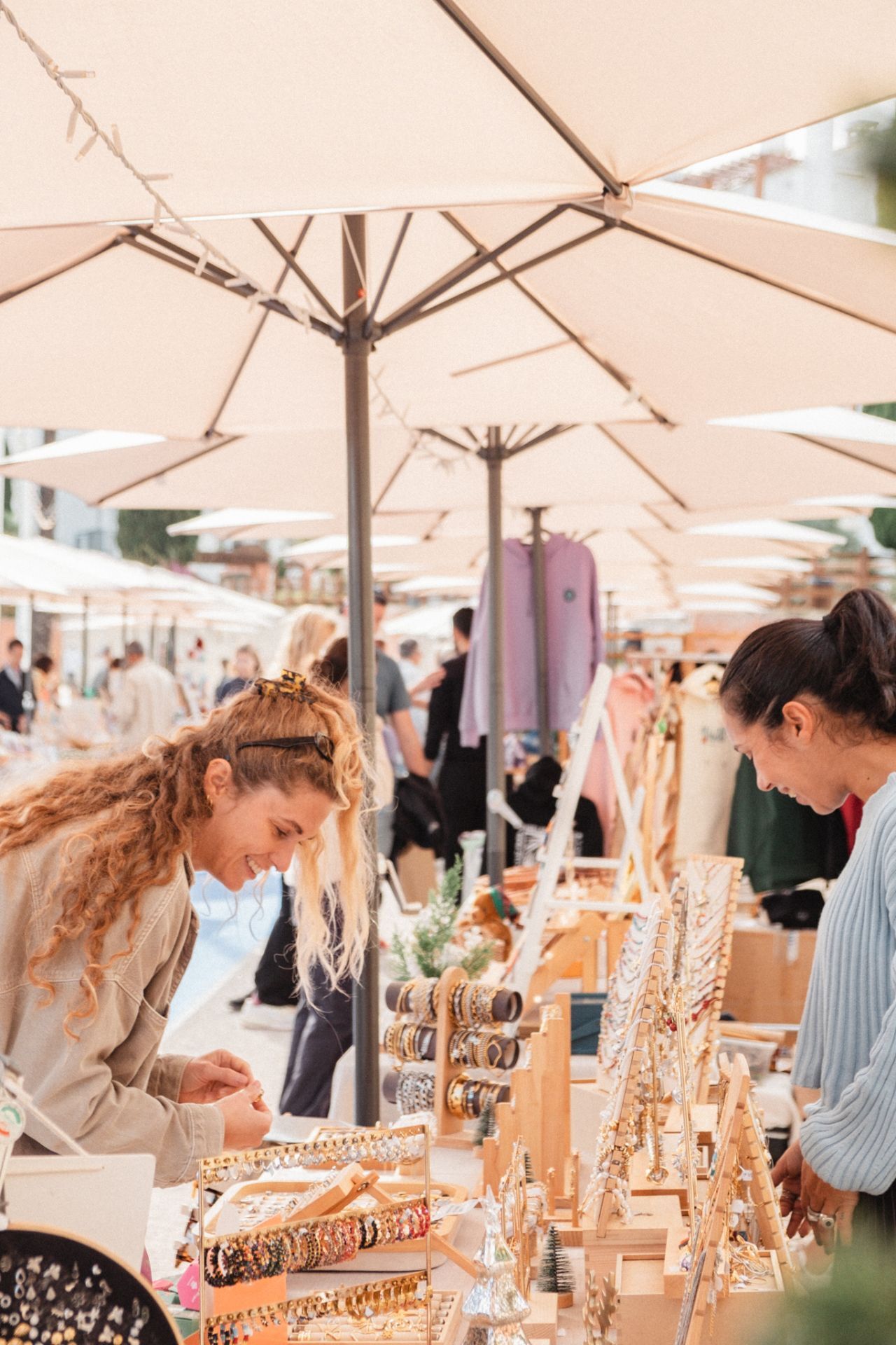 Vibrant outdoor market scene in Vilamoura, featuring two women browsing handcrafted jewelry under a row of white umbrellas, with a lively atmosphere in the background.