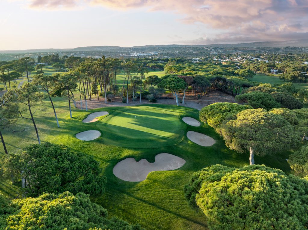 A breathtaking aerial view of the Old Course Vilamoura, showcasing its lush green fairways, well-placed sand bunkers, and iconic pine trees surrounded by the natural beauty of the Algarve landscape during golden hour.