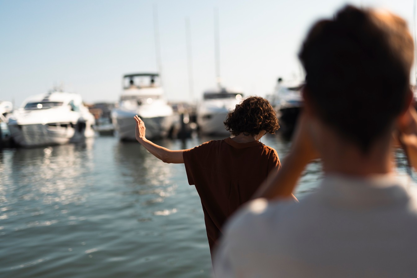 A person waving at a marina with luxury yachts docked in the background, under a clear sky.