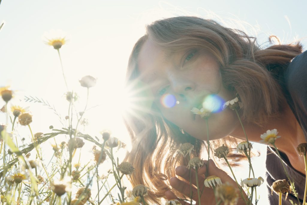 A young woman with wavy, light brown hair is captured in a sunlit field of daisies. The low-angle shot emphasizes her serene expression as she gently holds a daisy near her lips. Sunlight streams through, creating a lens flare effect that enhances the dreamy and natural atmosphere. The image conveys a strong connection with nature, tranquility, and an eco-friendly lifestyle.