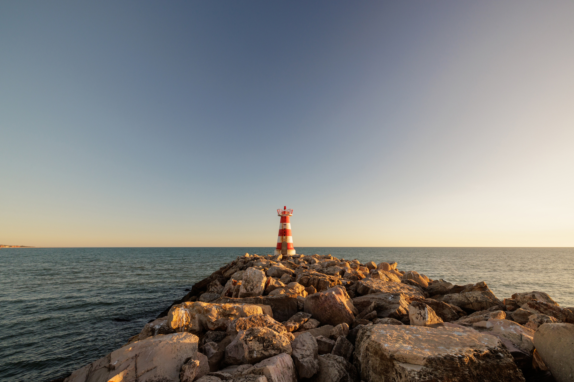 A small red and white striped lighthouse situated on a rocky breakwater extending into the sea. The sky is clear with a gradient from light blue near the horizon to darker blue at the top of the image. The sunlight casts warm tones on the rocks, indicating it might be either early morning or late afternoon.